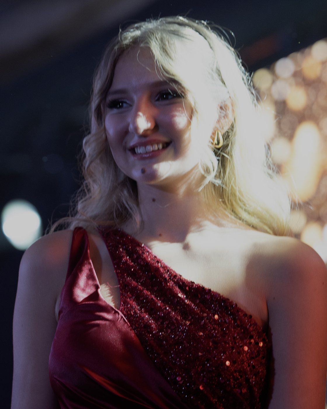 Léna Fiba souriante en robe bordeaux asymétrique à sequins, sur scène devant des lumières dorées