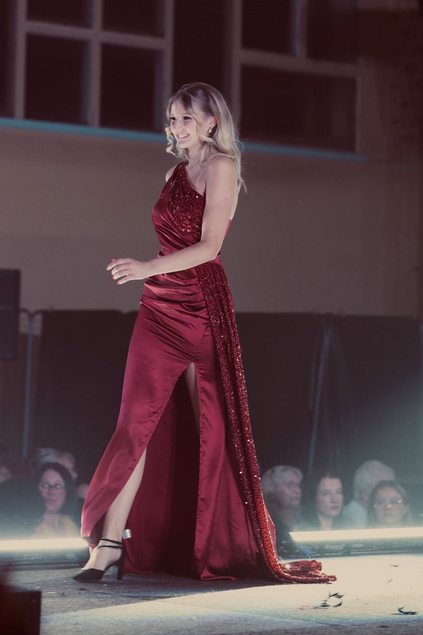 Léna Fiba défilant en longue robe bordeaux fendue à traîne pailletée, devant le public